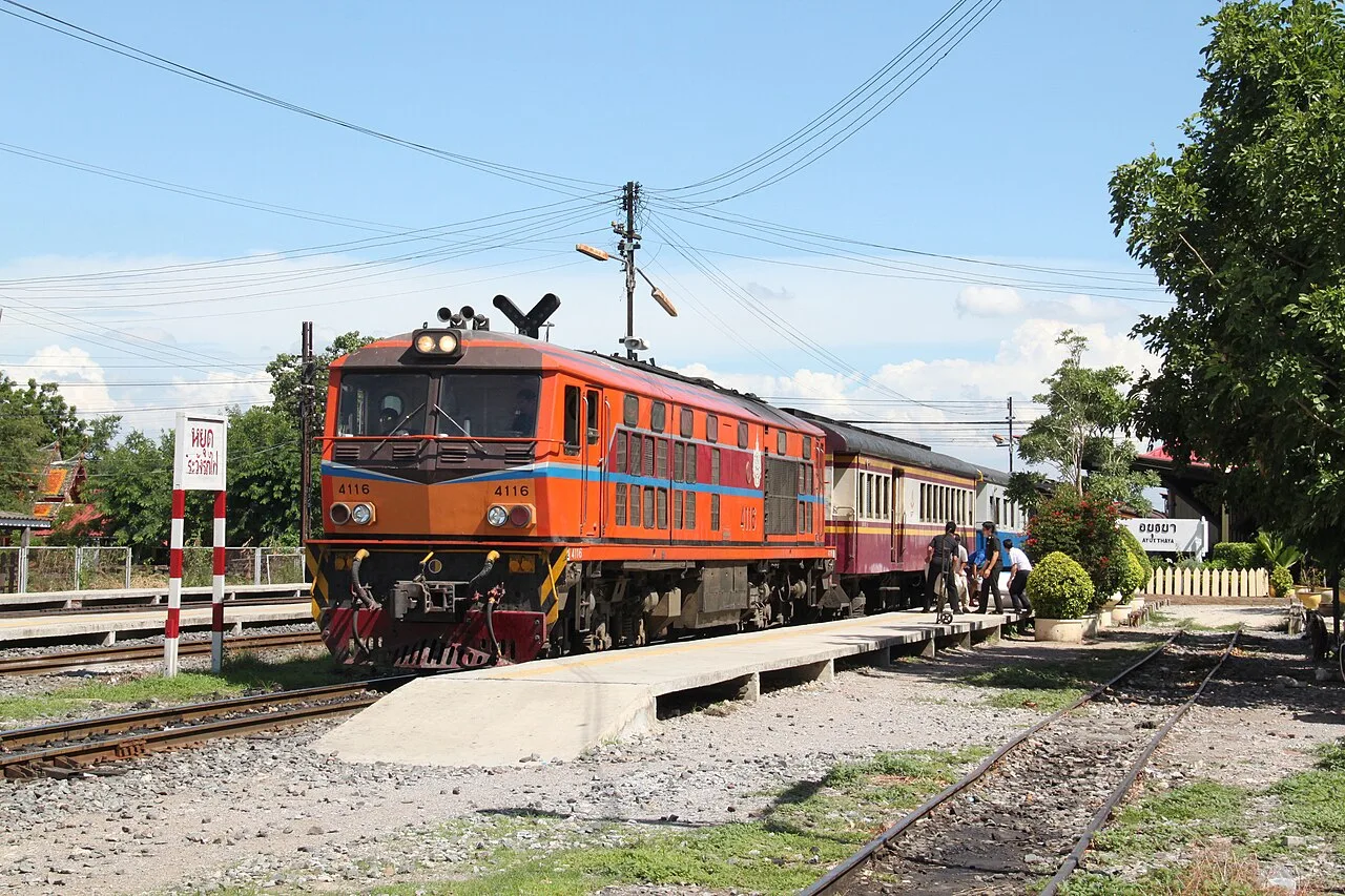 A diesel locomotive and passenger carriages standing at a platform of a small railway station on a sunny day, with signal lines overhead.